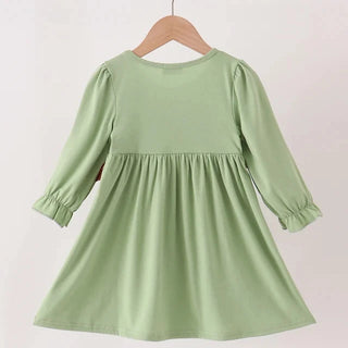A light sage green children's dress with three-quarter sleeves and a gathered waist, hanging on a wooden hanger from a wooden clothes rack against a plain off-white wall.