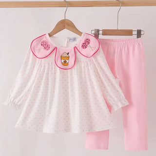 A children's two-piece outfit hanging on wooden hangers against a plain light background. The set includes a white long-sleeve top with tiny pink polka dots, a pink collar trimmed in pink, floral embroidery on the collar, and a small cupcake applique at the center, paired with solid light pink pants.