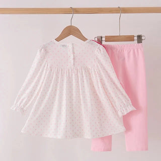 A children's two-piece outfit hanging on wooden hangers from a wooden clothes rack. The set includes a white long-sleeve top with tiny pink polka dots and a matching solid pink pair of pants.