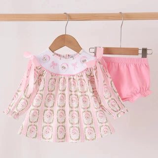 A baby girl's two-piece outfit hanging on wooden hangers against a plain light background. The set includes a long-sleeve dress with a pink and white floral pattern, a white collar with pink bows, and matching pink bloomers.