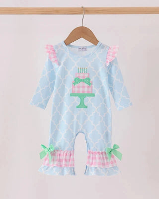 A baby girl's light blue jumpsuit with a pink and white trellis pattern, featuring a pink and white checkered cake design on the chest, pink ruffled shoulder straps, and pink ruffled leg bands with mint green bows, hanging on a wooden hanger.