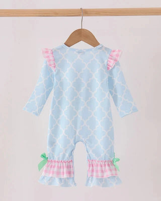 A baby romper in light blue with a white trellis pattern, featuring pink and white gingham ruffle details on the shoulders and legs, decorated with small green bows, hanging on a wooden hanger.