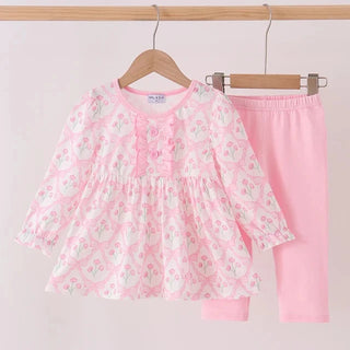 A two-piece girl's outfit set hanging on wooden hangers against a light pink background. The set includes a long-sleeve smocked blouse in white with a pink and green floral pattern, and solid light pink pants.