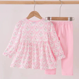 A toddler girl's two-piece clothing set hanging on wooden hangers from a wooden rod. The set includes a long-sleeve tunic top in light pink with a white and light green floral pattern, and matching solid light pink pants.