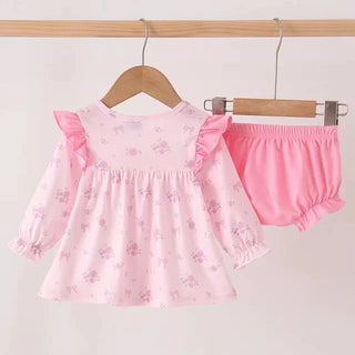A baby girl's two-piece outfit set consisting of a light pink long-sleeve dress with a subtle floral print and ruffle details, paired with matching solid pink ruffle bloomers, displayed on wooden hangers against a light background.