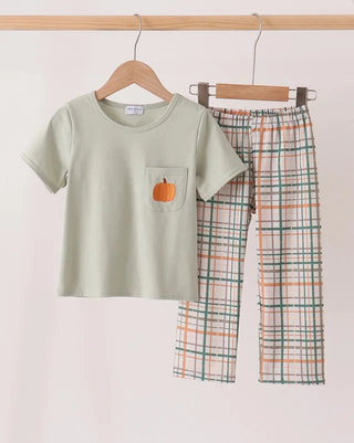 A children's two-piece outfit hanging on wooden hangers against a plain light background. The set includes a short-sleeve sage green top with a small orange pumpkin patch on the chest pocket, and matching plaid pants in sage green, orange, and white.