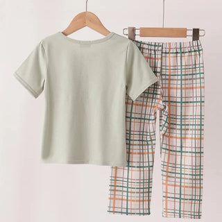 A two-piece children's pajama set hanging on wooden hangers from a light wood rod. The set includes a short-sleeve crewneck top in a pale sage green and matching plaid pants with a white base and orange, green, and blue plaid lines.