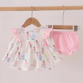 A baby girl's two-piece outfit hanging on wooden hangers against a light pink wall. The set includes a white ruffled dress with a pastel multicolor print of cartoon babies and strollers, and a pair of pink bloomers.