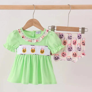A baby girl's two-piece Halloween outfit hanging on wooden hangers against a plain white wall. The set includes a light green short-sleeve dress with a white waistband decorated with yellow jack-o'-lanterns, and matching pink and purple skull-patterned shorts.