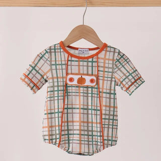 A baby short-sleeve romper hanging on a wooden hanger from a wooden rod. The romper features a green, orange, and white plaid pattern with a central orange pumpkin applique and orange trim.