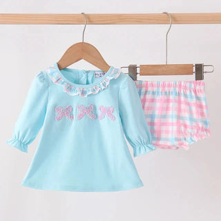 A toddler girl's two-piece clothing set hanging on wooden hangers against a plain white background. The set includes a light blue long-sleeve top with pink bow embroidery and a pink and blue plaid pair of shorts.