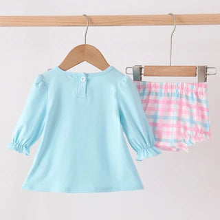 A toddler girl's two-piece clothing set hanging on wooden hangers from a wooden rod. The set includes a light blue long-sleeve top with ruffled cuffs and a matching pink and blue plaid diaper cover.