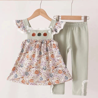 A girl's two-piece outfit set hanging on wooden hangers against a plain light background. The set includes a sleeveless dress with ruffled shoulders, a white base, and an all-over autumn pumpkin print in orange, beige, and gray, paired with sage green leggings featuring white lace trim at the cuffs.
