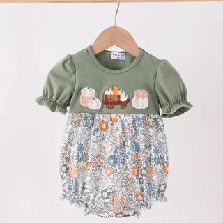 A toddler girl's short-sleeve romper top hanging on a wooden hanger against a plain white wall. The top features an olive green upper section with embroidered pumpkins and a blue, orange, and white floral patterned lower section.