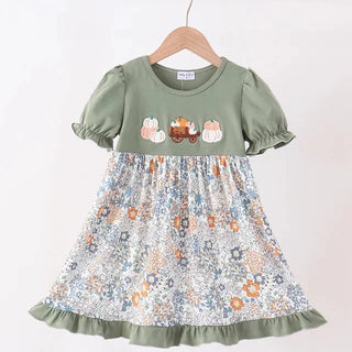 A toddler girl's dress hanging on a wooden hanger against a plain white wall. The dress has an olive green bodice with short puff sleeves and three embroidered pumpkins on the chest, and a matching olive green ruffled hem.