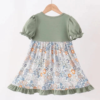 A toddler girl's dress with an olive green bodice and short puff sleeves, featuring a gathered floral skirt in blue, orange, and white. The dress is shown hanging on a wooden hanger from a light wood rod against a plain white background.