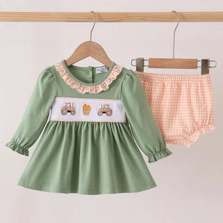 A baby girl's two-piece outfit set hanging on wooden hangers against a plain light background. The set includes a sage green long-sleeve dress with a ruffled collar, a white chest panel with embroidered tractors and cupcakes, and matching peach-and-white gingham bloomers.