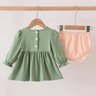 A toddler girl's two-piece clothing set hanging on wooden hangers from a wooden rod. The set includes a sage green long-sleeve peplum dress with white buttons and matching peach and white gingham bloomers.