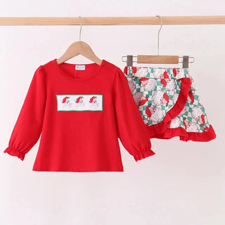 A children's two-piece outfit hanging on wooden hangers from a wooden rod. The set includes a bright red long-sleeve top with a small rectangular chest graphic and matching red-trimmed shorts with a green, white, and red floral print.