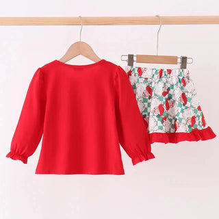 A two-piece children's outfit hanging on wooden hangers from a light wood rod. The set includes a bright red long-sleeve top with gathered cuffs and a matching patterned skirt with a red and green floral print on a white background and a red ruffled hem.