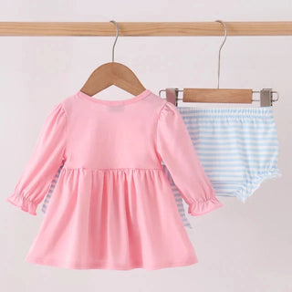 A baby girl's two-piece outfit hanging on wooden hangers from a light wood rod. The set includes a pink long-sleeve peplum dress with ruffled cuffs and matching light blue and white striped bloomers.