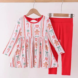 A children's two-piece outfit hanging on wooden hangers against a plain light background. The set includes a long-sleeve white dress with a festive gingerbread-themed print in red, green, and brown, paired with solid bright red leggings.