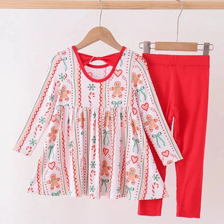 A toddler girl's two-piece outfit set consisting of a long-sleeve white top with a festive gingerbread and red heart pattern, paired with solid bright red leggings, hanging on wooden hangers against a plain white wall.