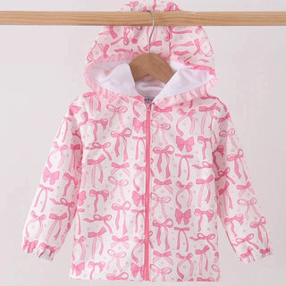 A children's hooded jacket with a pink and white bow pattern, hanging on a wooden hanger from a wooden clothes rack against a plain light background.