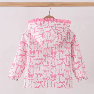 A children's hooded jacket with a pink and white bow pattern, hanging on a wooden hanger from a light wood clothing rack against a plain light background.