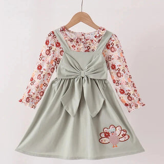 A toddler girl's dress hanging on a wooden hanger against a plain white wall. The dress features a sage green pinafore-style skirt with a large front bow, a long-sleeve floral shirt underneath, and a small embroidered turkey motif near the lower right hem.