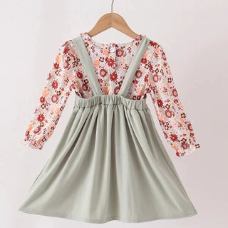 A toddler girl's dress hanging on a wooden hanger against a plain light background. The dress features a sage green overall-style skirt with shoulder straps and a long-sleeve floral insert top in red, orange, and cream tones.