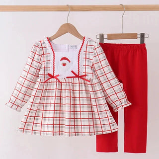 A toddler girl's two-piece clothing set hanging on wooden hangers against a plain white wall. The set includes a long-sleeve plaid dress in white, red, and light beige, with a white chest panel featuring a red embroidered Santa hat, and solid red pants.