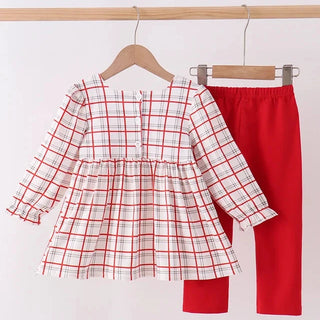 A toddler girl's two-piece outfit hanging on wooden hangers against a plain white wall. The set includes a long-sleeve plaid top in white with red and light gray lines, and solid bright red pants.