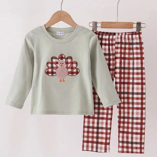 A children's two-piece pants set hanging on wooden hangers against a plain light background. The set includes a long-sleeve light sage green top with a turkey graphic on the front, and matching plaid pants in red, white, and brown.