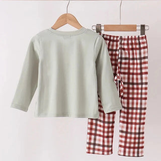 A children's two-piece outfit hanging on wooden hangers against a plain light background. The set includes a long-sleeve top in pale sage green and matching plaid pants in red, white, and dark brown.
