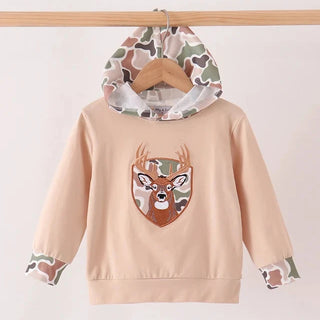 A children's beige hoodie sweatshirt hanging on a wooden hanger from a wooden rod. The hoodie features a camouflage pattern hood lining, camouflage patterned cuffs, and a central embroidered shield patch depicting a deer head with camouflage ears and face.