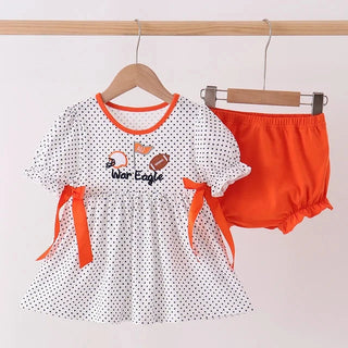 A baby girl's two-piece outfit hanging on wooden hangers against a light pink wall. The set includes a white short-sleeve top with black polka dots, orange trim, and orange side bows, featuring a 'War Eagle' football graphic on the chest, paired with bright orange bloomers.