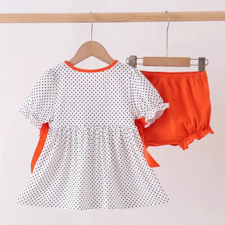 A toddler girl's two-piece outfit set hanging on wooden hangers from a wooden clothes rack. The set includes a white short-sleeve dress with black polka dots, orange trim at the collar, and orange side bows, paired with bright orange bloomers.