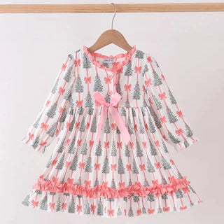 A long-sleeve tiered dress for toddlers, featuring a white base with a pattern of gray Christmas trees and pink bows, hanging on a wooden hanger against a plain light background.