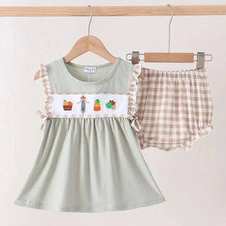 A two-piece baby girl's fall outfit set hanging on wooden hangers against a light beige wall. The set includes a sage green sleeveless dress with a white chest panel featuring colorful autumn-themed illustrations (pumpkin, acorn, scarecrow) and ruffle trim, paired with beige and white gingham check shorts with ruffle trim.