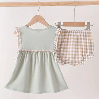 A two-piece baby girl's outfit hanging on wooden hangers from a light wood rod. The set includes a sage green sleeveless dress with beige and white ruffle trim on the shoulders and waist, and matching beige and white gingham check bloomers with ruffle trim.