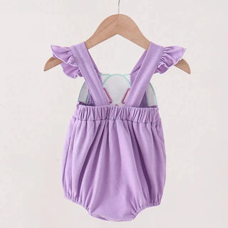 A baby romper in light purple with ruffled shoulder straps, hanging on a wooden hanger from a wooden clothes rack against a plain off-white background.