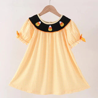 A toddler girl's short-sleeve dress with a yellow and white gingham pattern, featuring a black collar decorated with three candy corn appliqués, hanging on a wooden hanger against a plain light background.