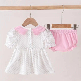 A baby girl's two-piece outfit set hanging on wooden hangers from a light wood rod. The set includes a white short-sleeve dress with tiny black polka dots and a pink Peter Pan collar, paired with matching pink bloomers.