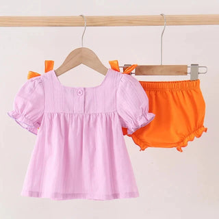 A baby girl's two-piece outfit hanging on wooden hangers against a plain light background. The set includes a light pink short-sleeve smocked blouse with orange shoulder bows and matching orange ruffled bloomers.