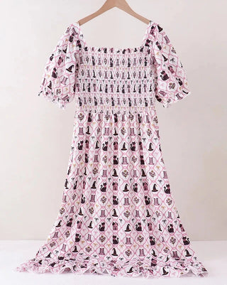 A pink and white smocked dress with a black cat pattern, hanging on a wooden hanger against a light beige wall.