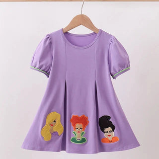 A lavender purple short-sleeve dress with puff sleeves, hanging on a wooden hanger. The dress features three cartoon character face patches on the front hem and light green piping along the sleeve cuffs.