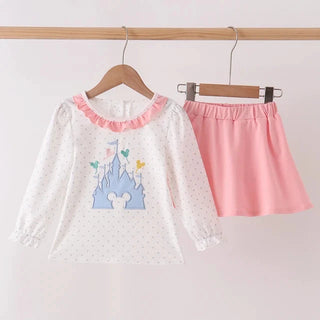 A children's two-piece outfit hanging on wooden hangers against a plain light background. The set includes a white long-sleeve blouse with a pink ruffled collar, tiny white polka dots, and a blue castle graphic on the front, paired with a solid pink skirt.