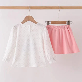 A two-piece girls' outfit set hanging on wooden hangers from a light wood rod. The set includes a white long-sleeve blouse with tiny pink polka dots and a pink elastic-waist mini skirt.