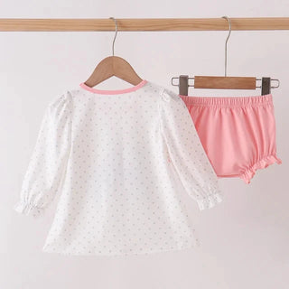 A two-piece baby clothing set hanging on wooden hangers from a wooden rod. The set includes a white long-sleeve blouse with tiny pink polka dots and a pink collar, paired with pink elastic-waist bloomers with ruffled trim.